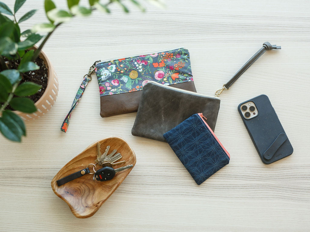 table with wristlet, coin pouch and leather wristlet. keys in wood dish with a plant a phone laying on the table