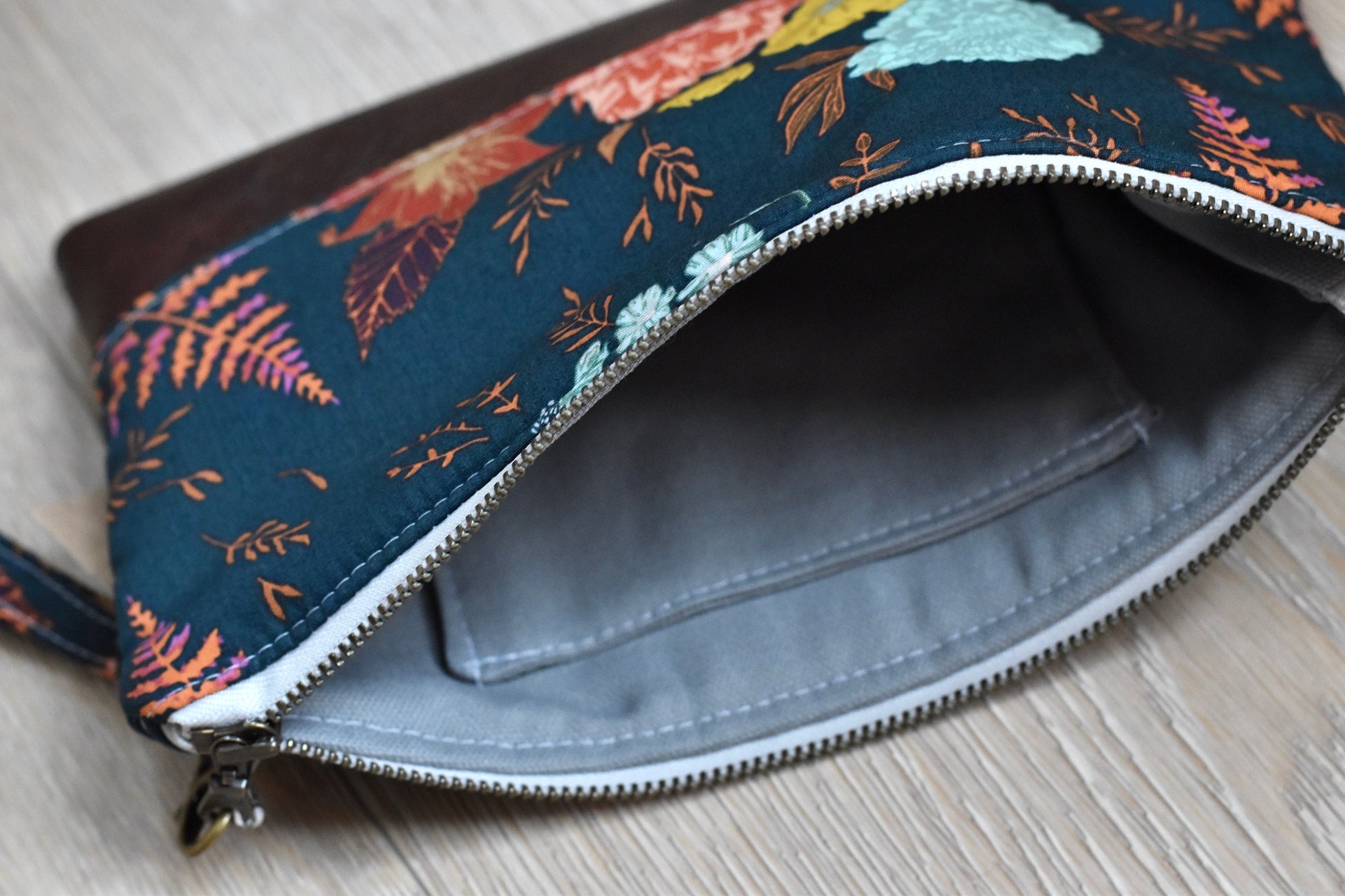 floral and fern navy wristlet with matching wrist strap and brown vinyl along the bottom. antique brass zipper. Lining is gray with slip pocket for cards