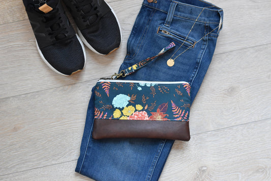 floral and fern navy wristlet with matching wrist strap and brown vinyl along the bottom. antique brass zipper. styled with jeans and black tennis shoes