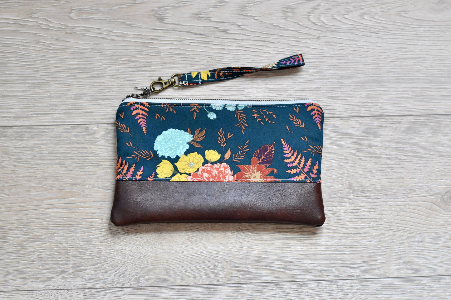 floral and fern navy wristlet with matching wrist strap and brown vinyl along the bottom. antique brass zipper. 