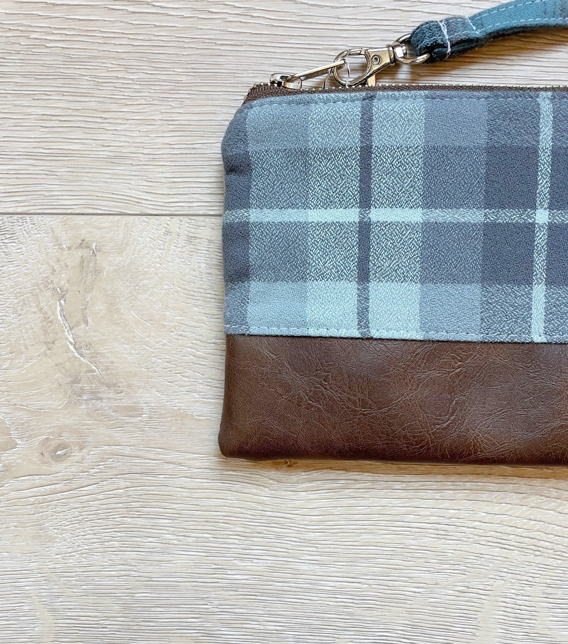 Blue and gray flannel wristlet with brown vinyl along the bottom, closed with a silver zipper. matching flannel wrist strap
