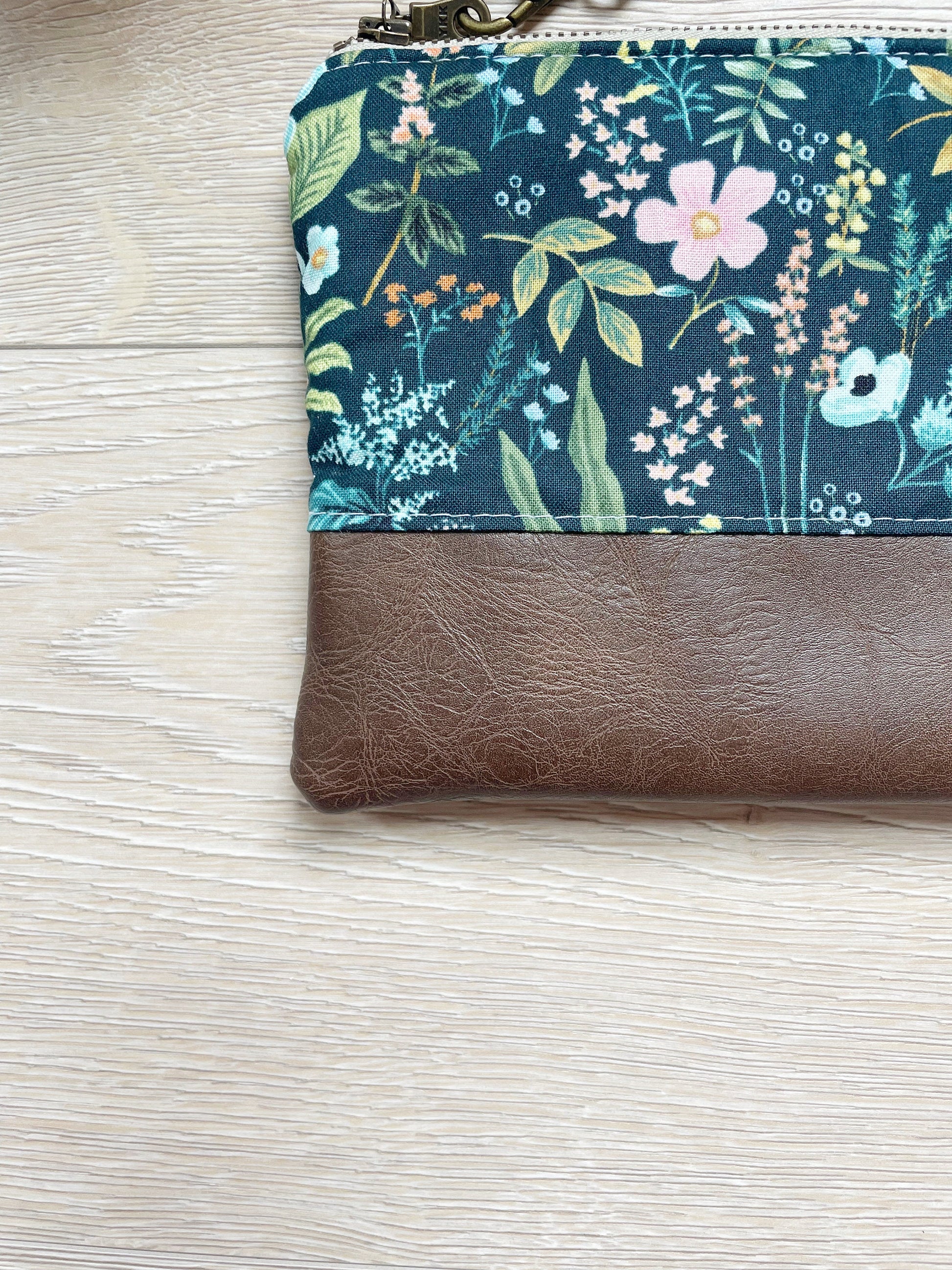 Blue floral wristlet with pink flowers and brown vinyl along the bottom. wristlet has matching wrist strap and antique brass zipper