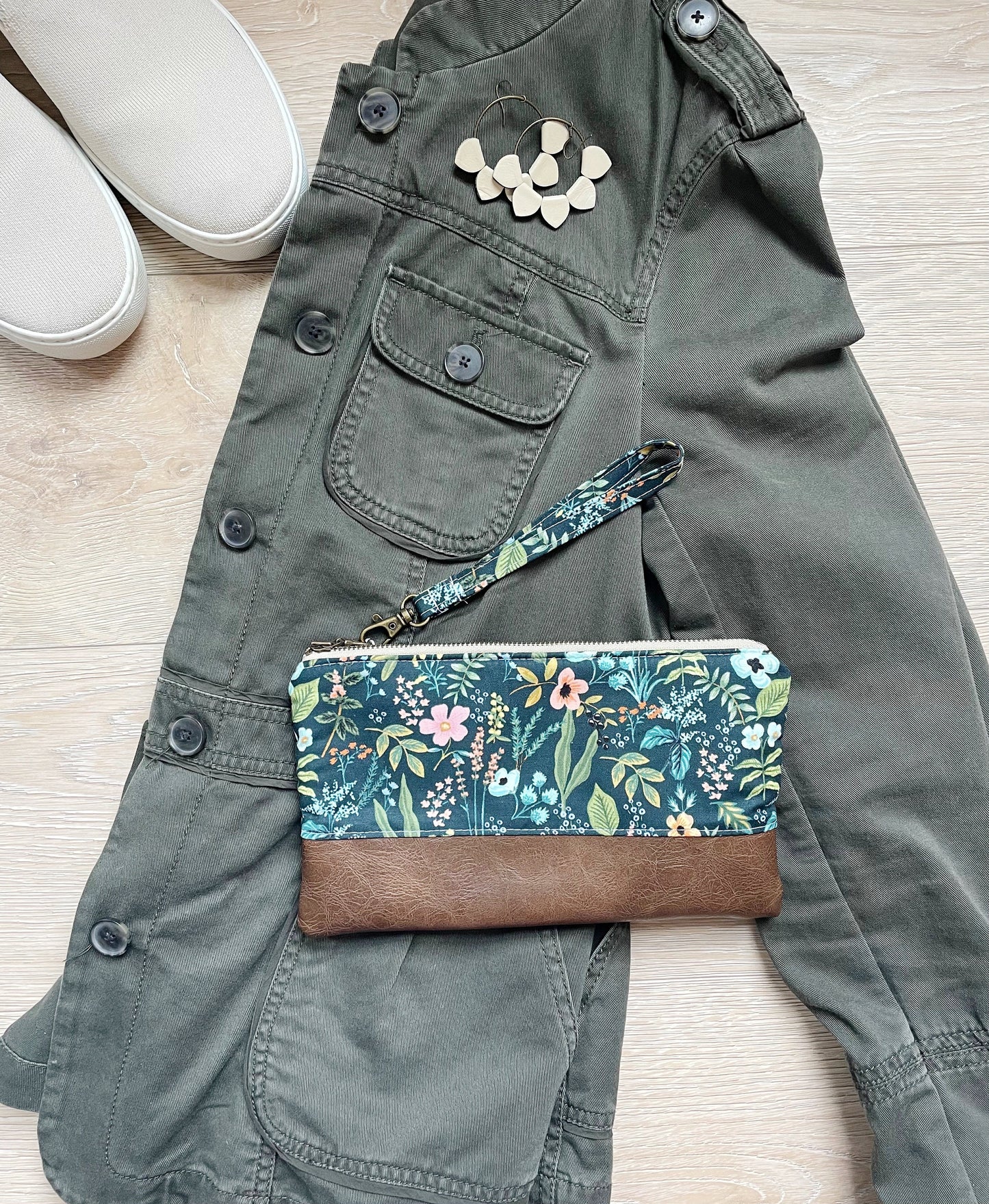 Blue floral wristlet with pink flowers and brown vinyl along the bottom. wristlet has matching wrist strap and antique brass zipper, styled with green coat