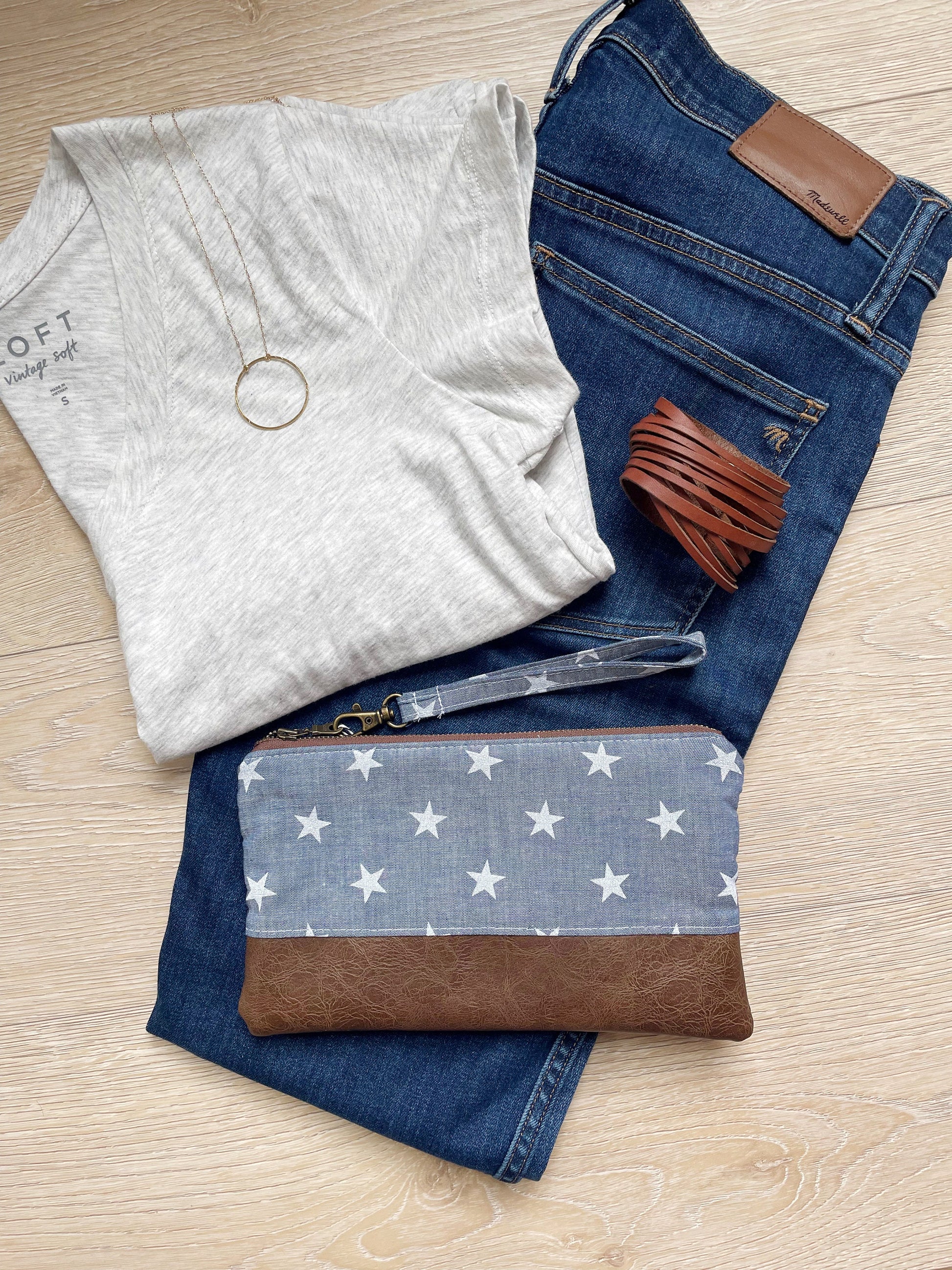chambray wristlet with white stars and matching wrist strap. brown vinyl along the bottom and antique brass zipper. styled with jeans and a gray shirt with leather cuff and gold necklace.